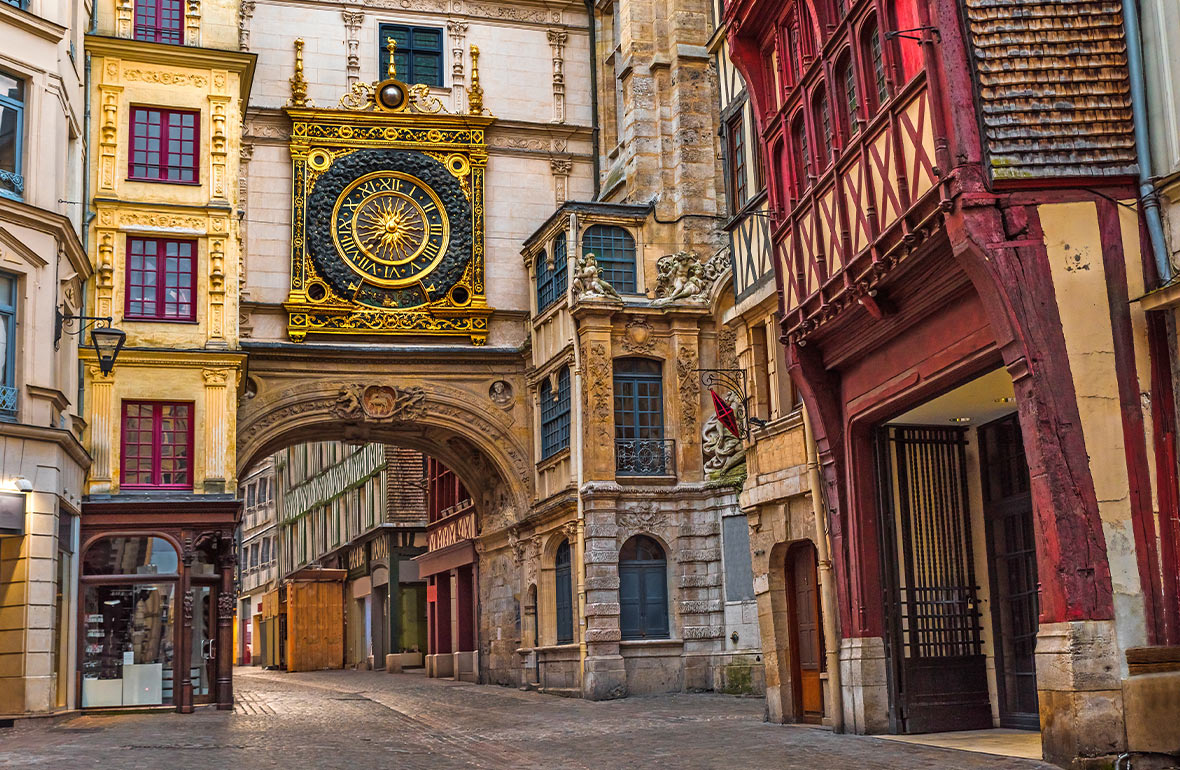 Large ornate golden clock above an archway on an old cobbled street lined with old buildings